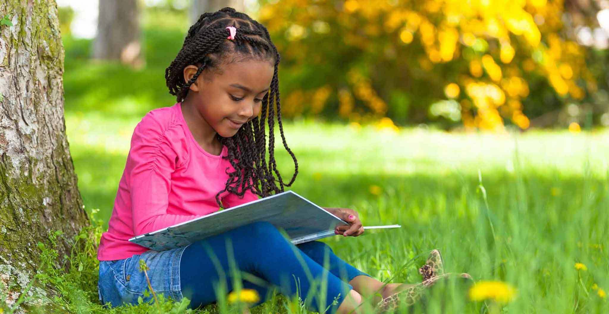 Child Reading Book Against a Tree in the Grass